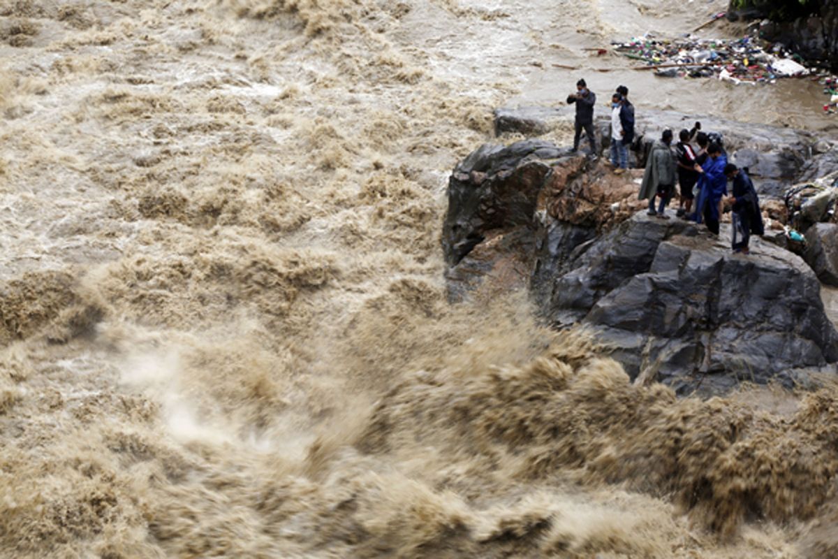A group of onlookers on a rocky ledge next to a violent muddy river torrent