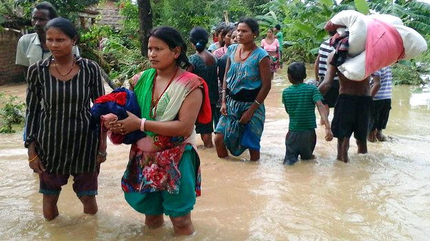 Women and children wading through knee-deep muddy floodwater carrying salvaged belongings